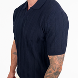 A man wearing a dark navy blue short-sleeve knit polo shirt and light beige pants, shown from the waist up against a plain white background.