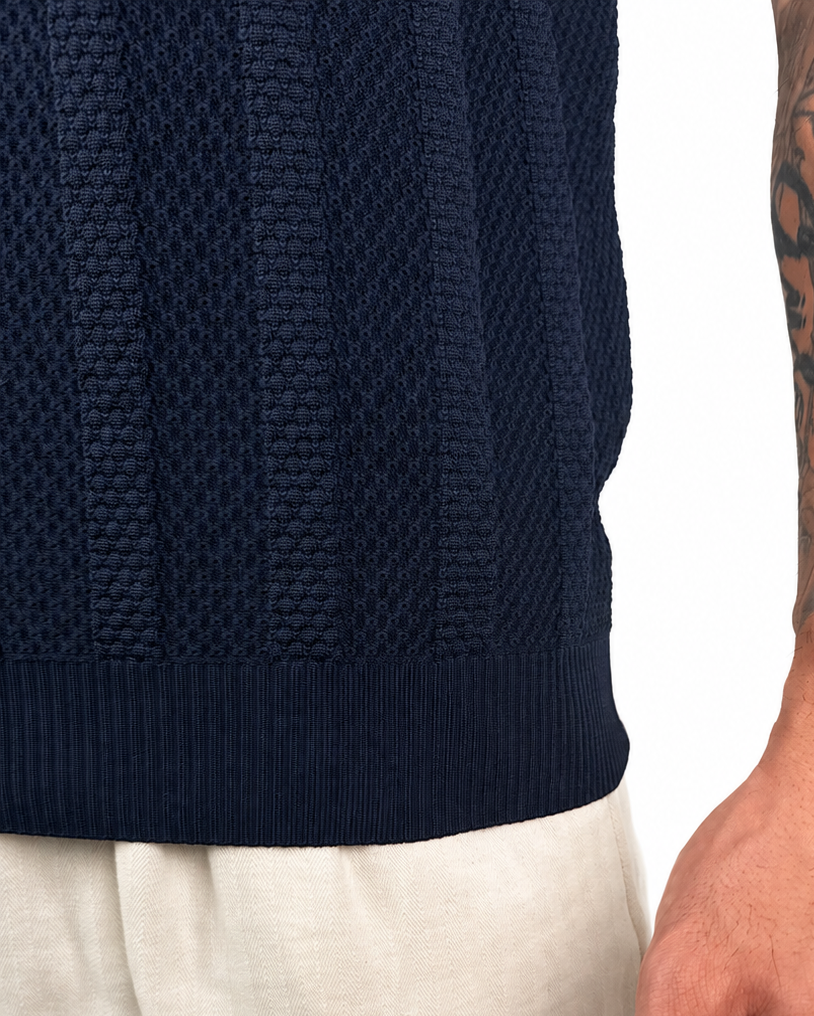 Close-up of a person wearing a dark navy ribbed knit sweater and light beige trousers, with the tattooed right arm and hand visible against a plain white background.