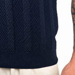 Close-up of a person wearing a dark navy ribbed knit sweater and light beige trousers, with the tattooed right arm and hand visible against a plain white background.
