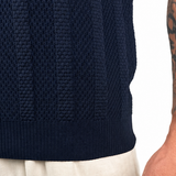 Close-up of a person wearing a dark navy ribbed knit sweater and light beige trousers, with the tattooed right arm and hand visible against a plain white background.