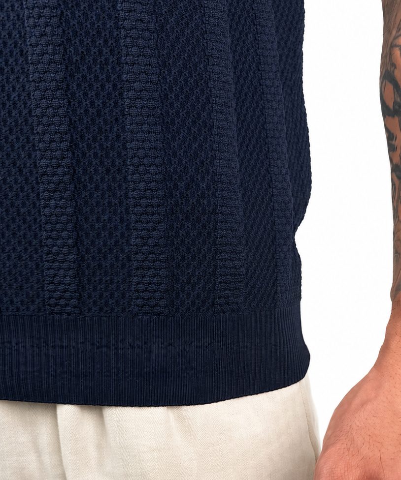 Close-up of a person wearing a dark navy ribbed knit sweater and light beige trousers, with the tattooed right arm and hand visible against a plain white background.