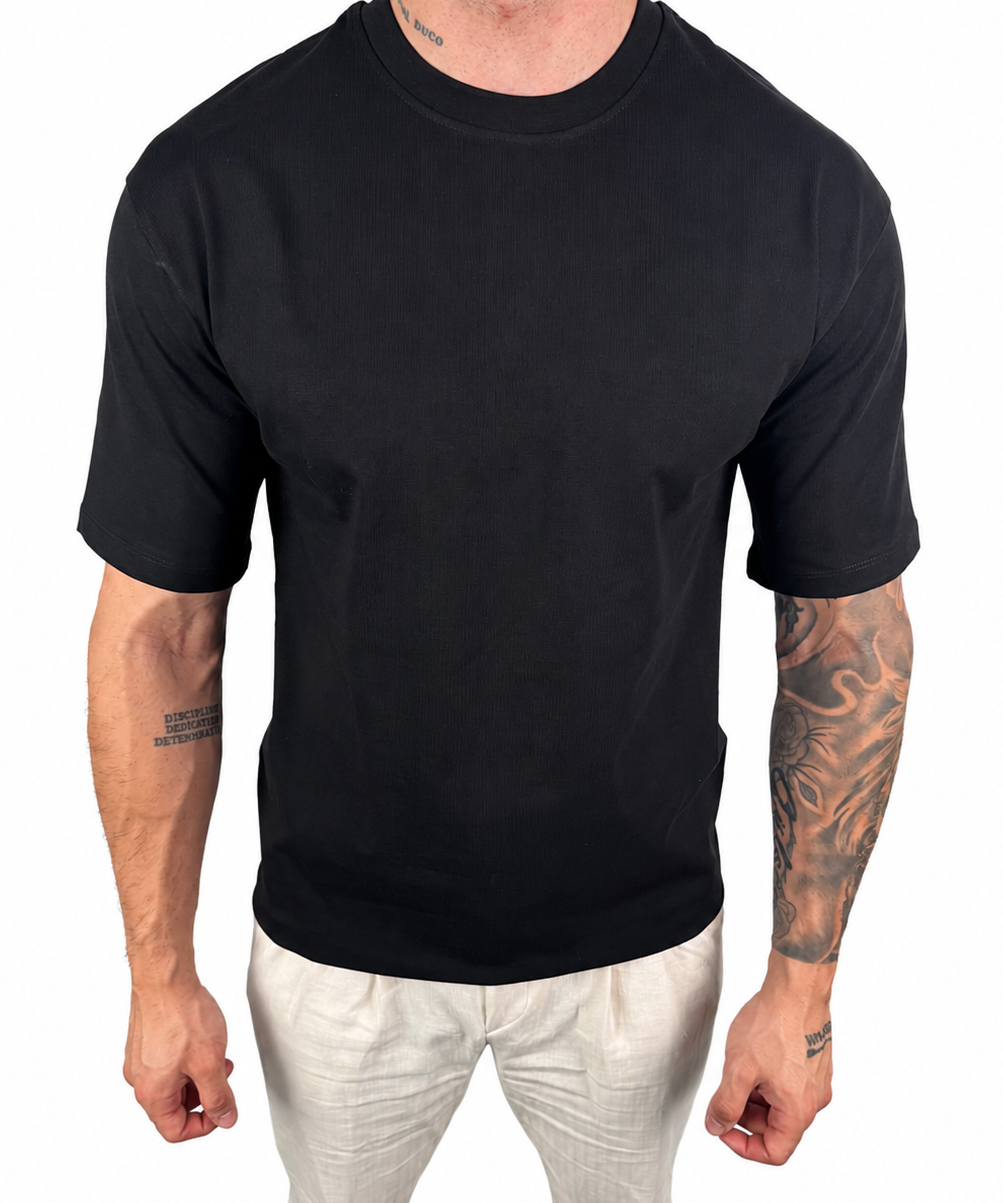 A man wearing a plain black short-sleeve crewneck t-shirt and off-white pants, shown from the neck down against a solid white background.