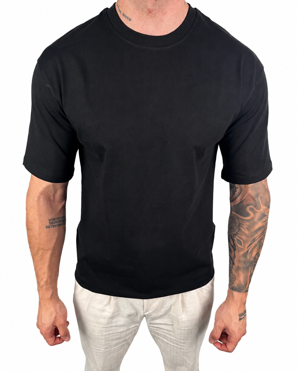 A man wearing a plain black short-sleeve crewneck t-shirt and off-white pants, shown from the neck down against a solid white background.