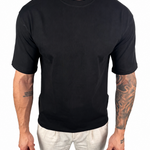A man wearing a plain black short-sleeve crewneck t-shirt and off-white pants, shown from the neck down against a solid white background.