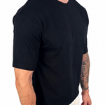 A man wearing a black short-sleeve crewneck t-shirt and off-white linen pants, shown from a side-back angle against a plain white background.