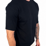 A man wearing a black short-sleeve crewneck t-shirt and off-white linen pants, shown from a side-back angle against a plain white background.