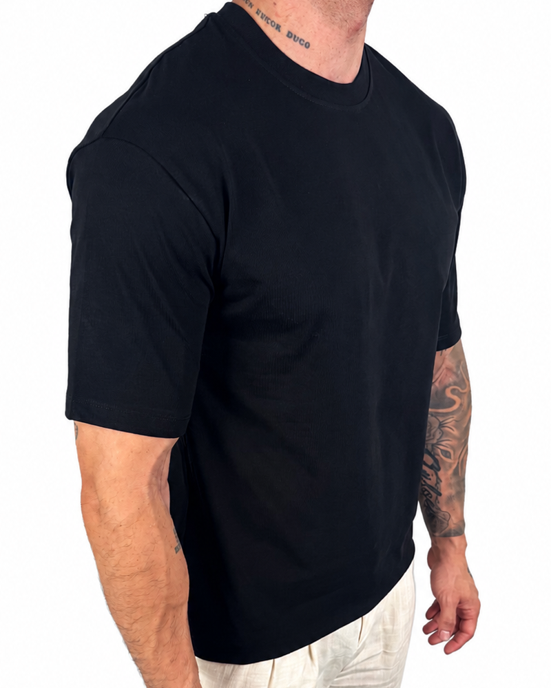 A man wearing a black short-sleeve crewneck t-shirt and off-white linen pants, shown from a side-back angle against a plain white background.