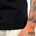 A close-up of a man's tattooed forearm and hand wearing a black t-shirt and light beige linen trousers, with a temporary tattoo on his wrist reading 'WARRIOR'.
