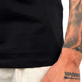 A close-up of a man's tattooed forearm and hand wearing a black t-shirt and light beige linen trousers, with a temporary tattoo on his wrist reading 'WARRIOR'.