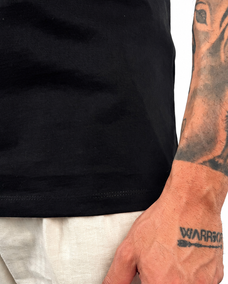 A close-up of a man's tattooed forearm and hand wearing a black t-shirt and light beige linen trousers, with a temporary tattoo on his wrist reading 'WARRIOR'.