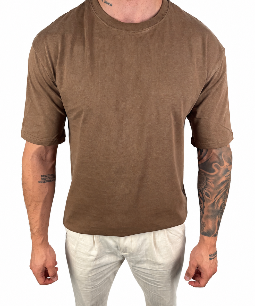 A man wearing a brown short-sleeve crewneck t-shirt and off-white pants, shown from the neck down against a plain white background.
