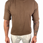 A man wearing a brown short-sleeve crewneck t-shirt and off-white pants, shown from the neck down against a plain white background.