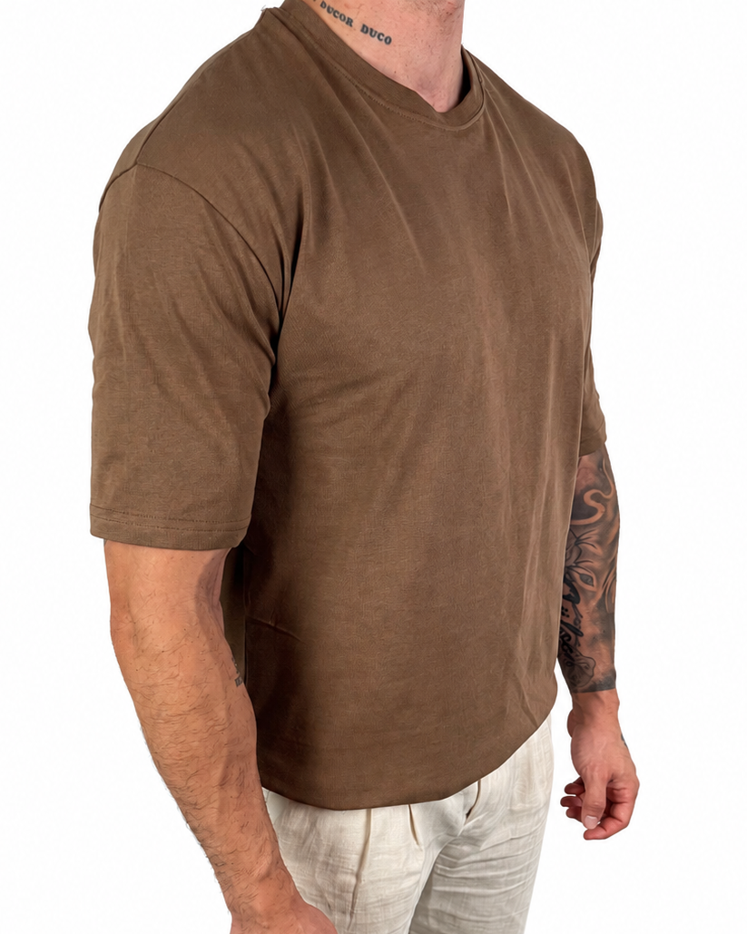 A man wearing a brown short-sleeve crewneck t-shirt and light beige linen pants, shown from the neck down against a solid white background.