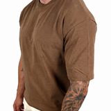 A man wearing a brown short-sleeve crewneck t-shirt and off-white pants, shown from the side against a plain white background.