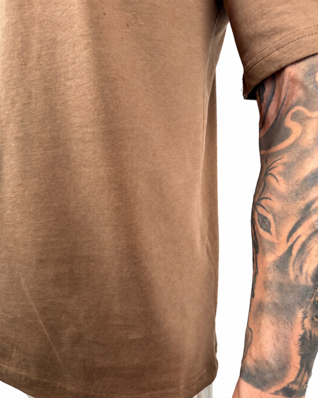 Close-up of a person wearing a brown short-sleeve t-shirt, showing the side and back of the garment with a tattooed arm visible against a white background.