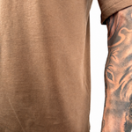 Close-up of a person wearing a brown short-sleeve t-shirt, showing the side and back of the garment with a tattooed arm visible against a white background.
