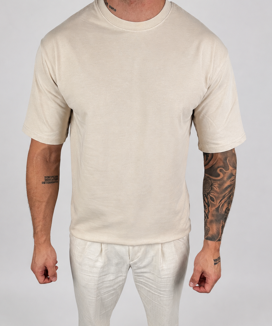 A man wearing a plain cream-colored short-sleeve crewneck t-shirt and matching cream pants, standing against a light gray studio background.