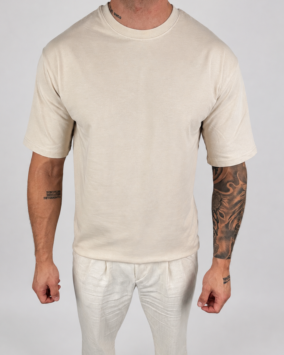 A man wearing a plain cream-colored short-sleeve crewneck t-shirt and matching cream pants, standing against a light gray studio background.