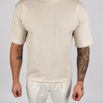 A man wearing a plain cream-colored short-sleeve crewneck t-shirt and matching cream pants, standing against a light gray studio background.
