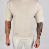 A man wearing a plain cream-colored short-sleeve crewneck t-shirt and matching cream pants, standing against a light gray studio background.