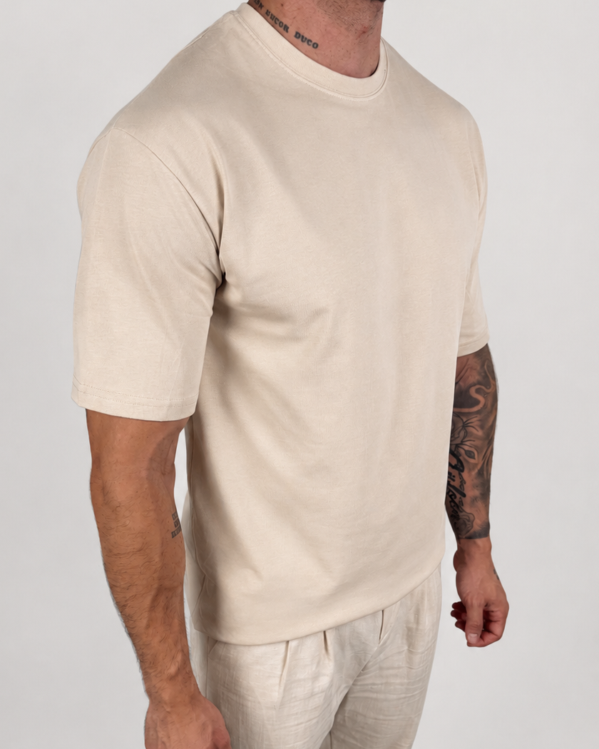 A man wearing a light beige short-sleeve crewneck t-shirt and matching light beige linen pants, standing against a plain light gray studio background.