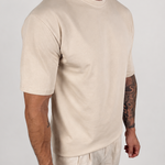 A man wearing a light beige short-sleeve crewneck t-shirt and matching light beige linen pants, standing against a plain light gray studio background.