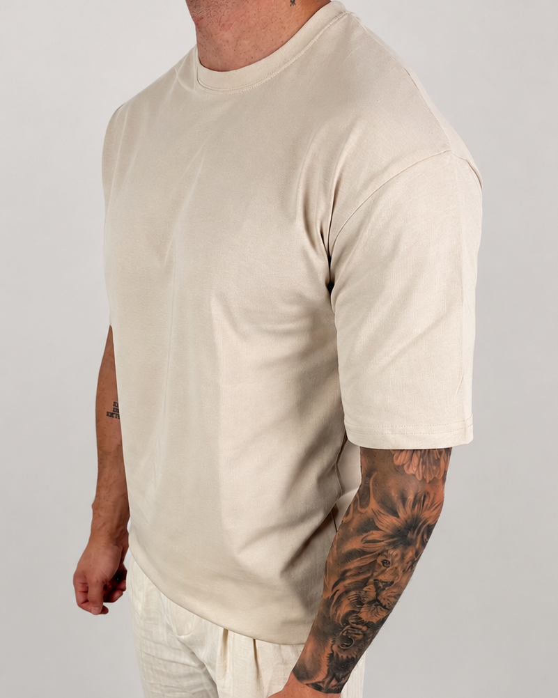 A man wearing a light beige crew-neck t-shirt and matching light beige pants, standing against a neutral light gray studio background.