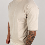 A man wearing a light beige crew-neck t-shirt and matching light beige pants, standing against a neutral light gray studio background.