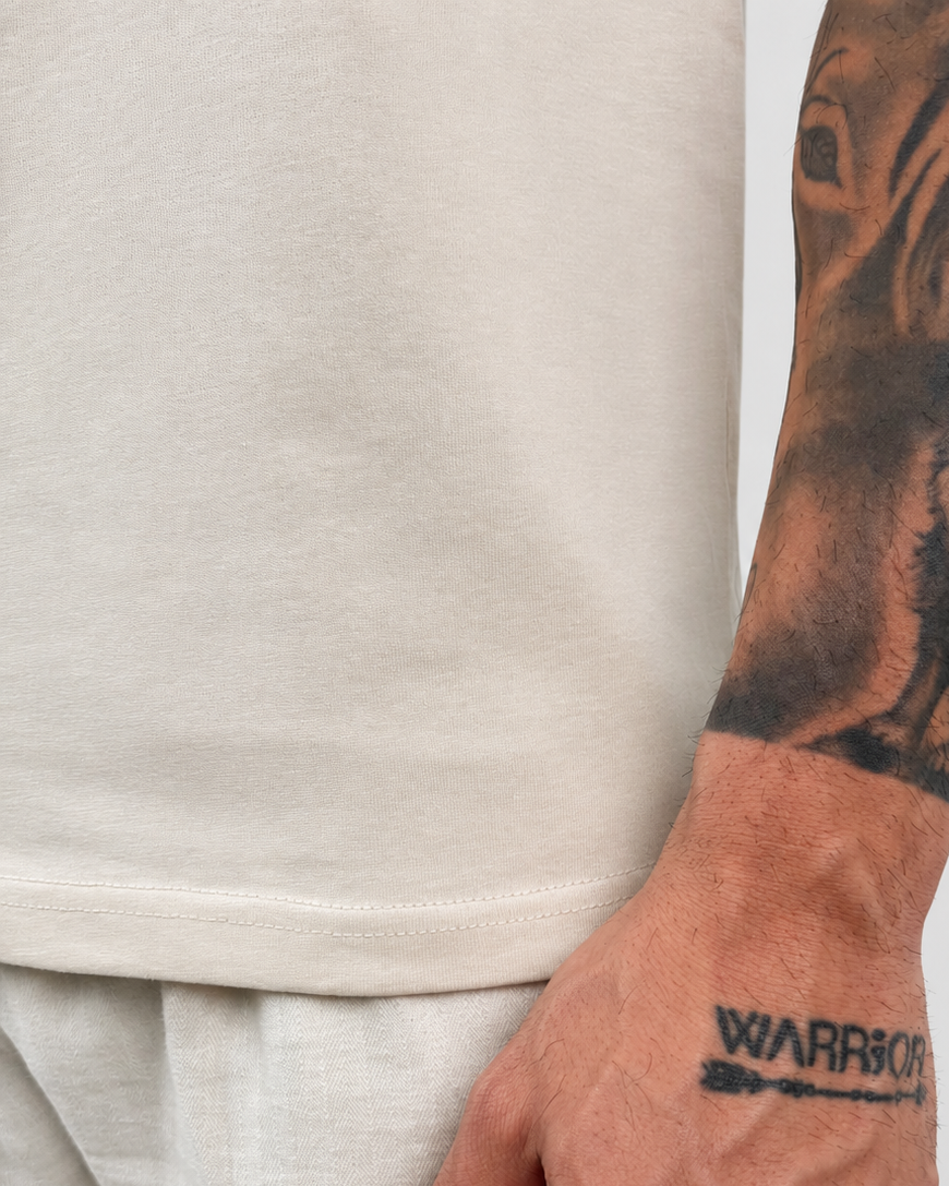 Close-up of a person wearing a light beige crewneck t-shirt and matching light beige trousers, with a detailed black tattoo of a dog on their forearm and 'WARRIOR' printed on their wrist.
