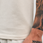 Close-up of a person wearing a light beige crewneck t-shirt and matching light beige trousers, with a detailed black tattoo of a dog on their forearm and 'WARRIOR' printed on their wrist.
