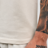 Close-up of a person wearing a light beige crewneck t-shirt and matching light beige trousers, with a detailed black tattoo of a dog on their forearm and 'WARRIOR' printed on their wrist.