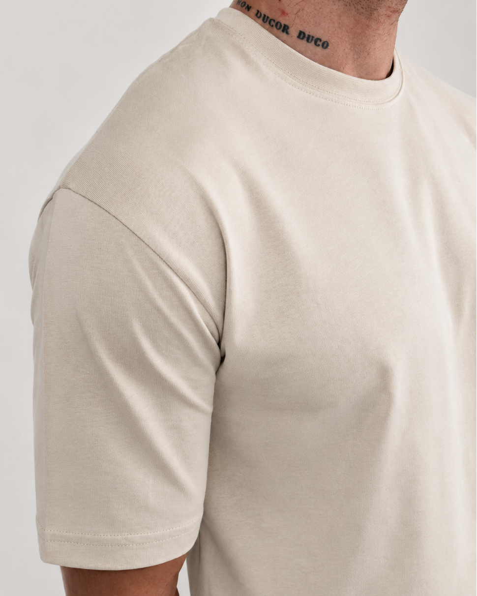 A close-up side view of a man wearing a light beige crew-neck t-shirt and matching light pants against a plain light gray background.