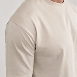 A close-up side view of a man wearing a light beige crew-neck t-shirt and matching light pants against a plain light gray background.