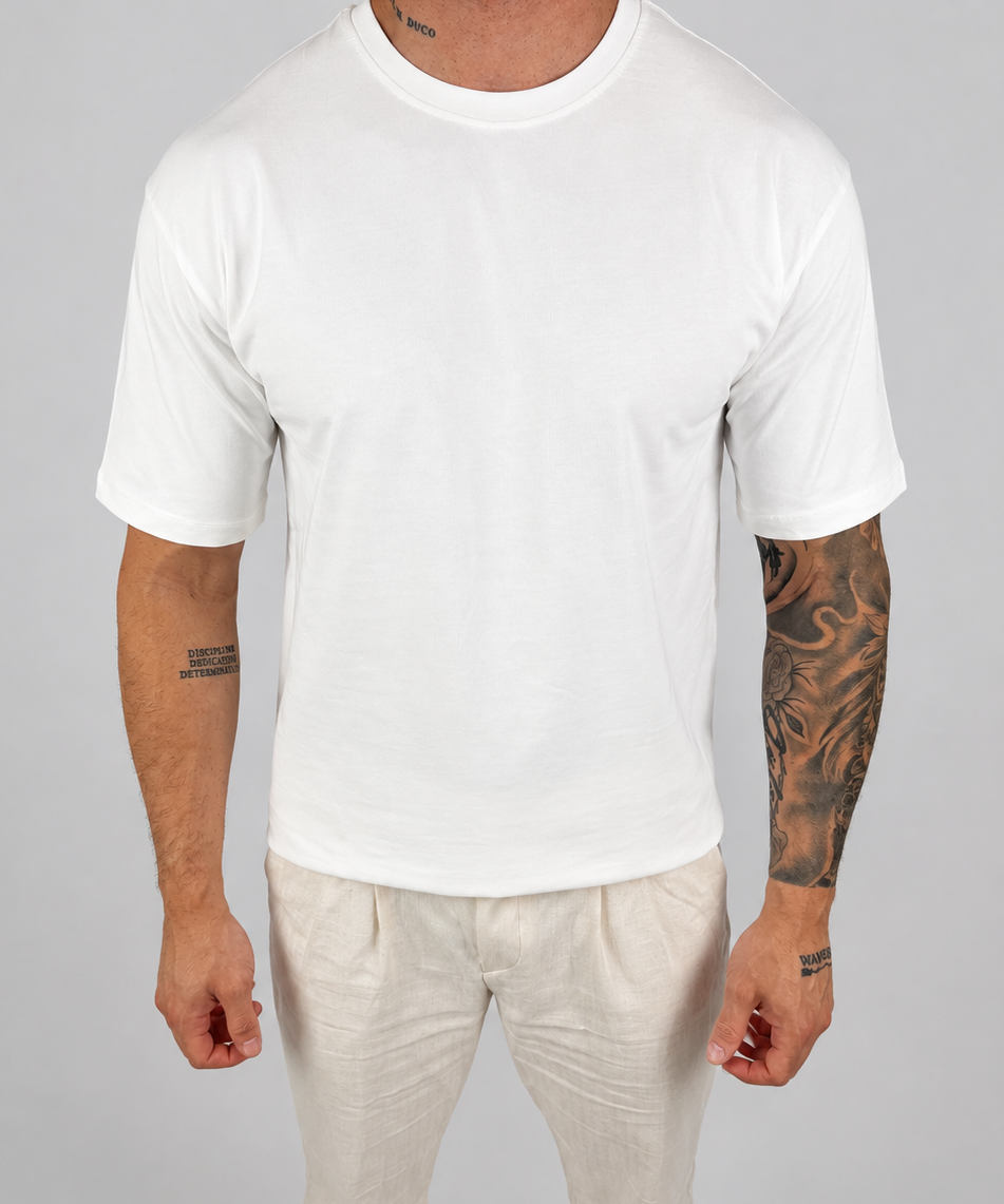A man wearing a plain white short-sleeve crewneck t-shirt and off-white trousers, standing against a neutral light gray background.