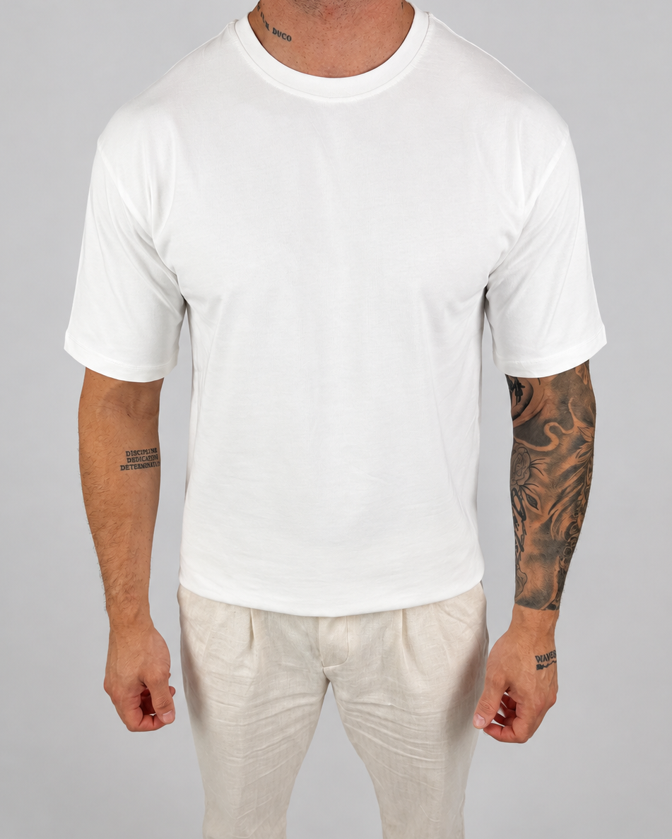 A man wearing a plain white short-sleeve crewneck t-shirt and off-white trousers, standing against a neutral light gray background.