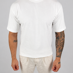 A man wearing a plain white short-sleeve crewneck t-shirt and off-white trousers, standing against a neutral light gray background.