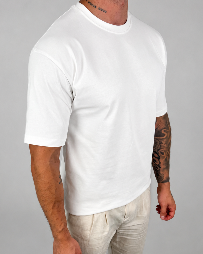 A man wearing a plain white short-sleeve crewneck t-shirt and light beige linen pants, standing against a neutral gray studio background.