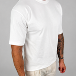 A man wearing a plain white short-sleeve crewneck t-shirt and light beige linen pants, standing against a neutral gray studio background.