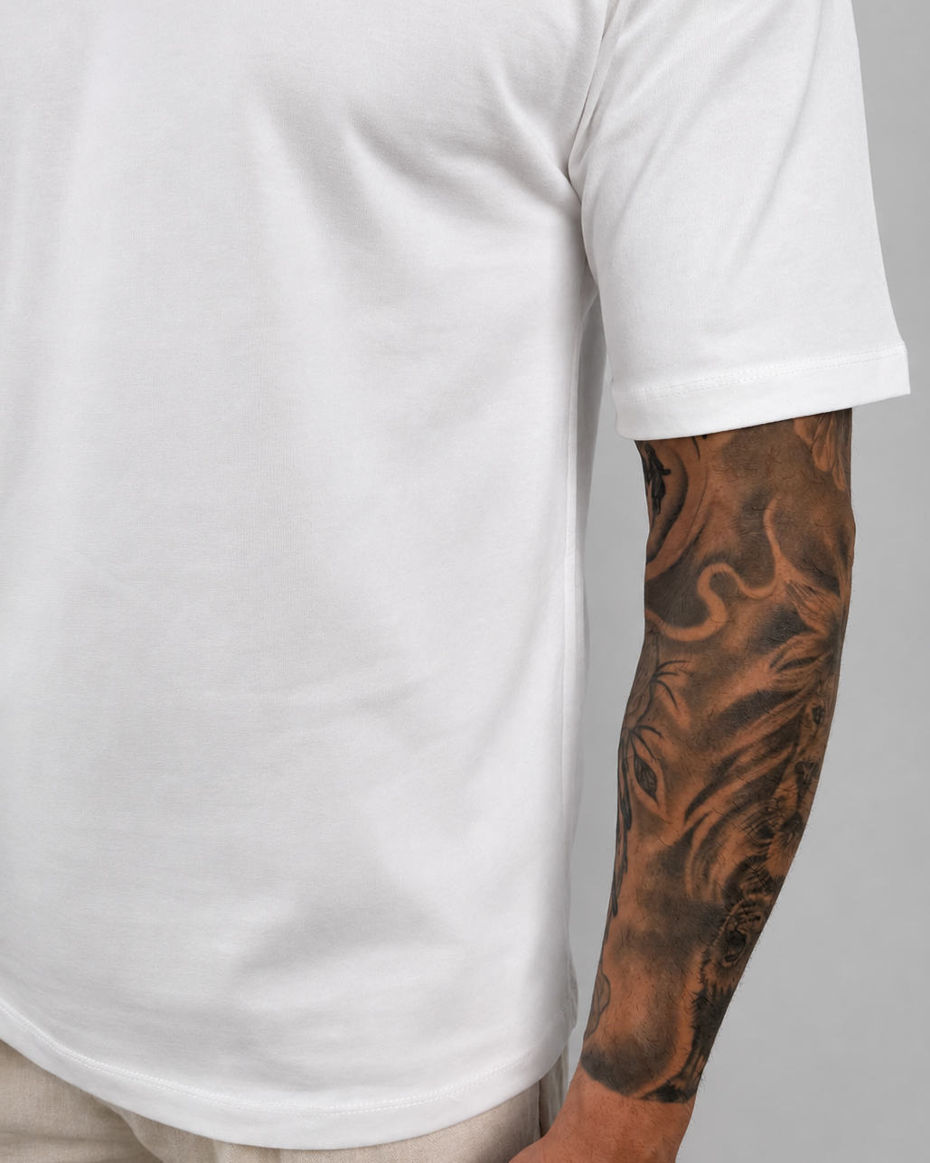 A close-up of a person wearing a plain white short-sleeve t-shirt and light beige trousers, showing a detailed black tattoo on their right arm against a neutral gray background.