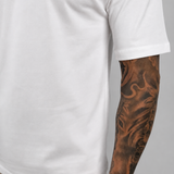 A close-up of a person wearing a plain white short-sleeve t-shirt and light beige trousers, showing a detailed black tattoo on their right arm against a neutral gray background.